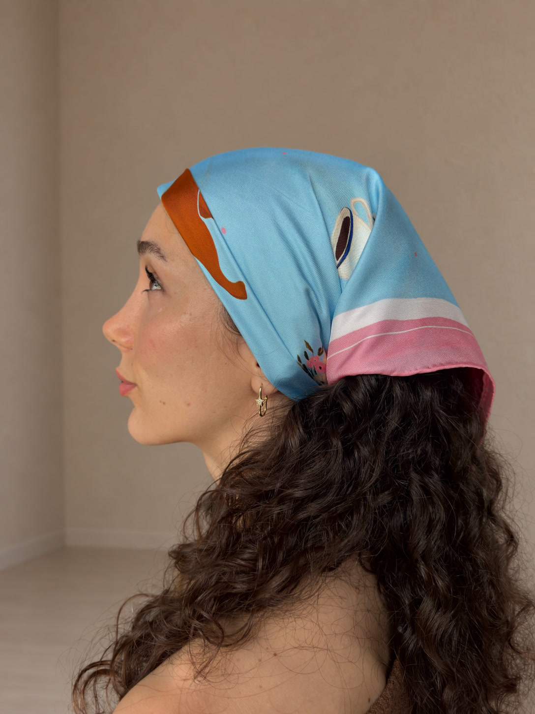 The sky-blue, pink and ivory Dachshund silk scarf worn over curly hair, seen in profile with the illustrated coffee cup and silk folds visible.
